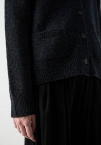 Gros plan sur une personne portant un cardigan en maille gris foncé avec des boutons à l'avant et une poche, associé à une jupe plissée noire.