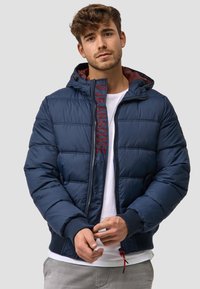 Chaqueta acolchada navy con capucha desmontable, con cierre frontal y logo bordado. Puños y dobladillo acanalados, textura acolchada y forro en contraste.