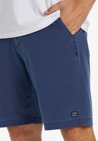 Marineblaue Shorts aus leichtem Stoff mit glatter Textur, seitlichen Taschen und einem kleinen schwarzen Logodetail am Saum.