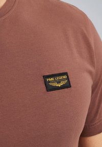 Braunes kurzärmliges T-Shirt aus weichem Stoff, mit einem schwarzen rechteckigen Patch mit gelber Schrift und Flügel-Logo im Brustbereich.