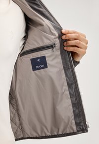 Person, der eine dunkle Lederjacke mit hellgrauem Innenfutter hält, das eine mit Reißverschluss versehenen Tasche und ein JOOP!-Markenschild zeigt.