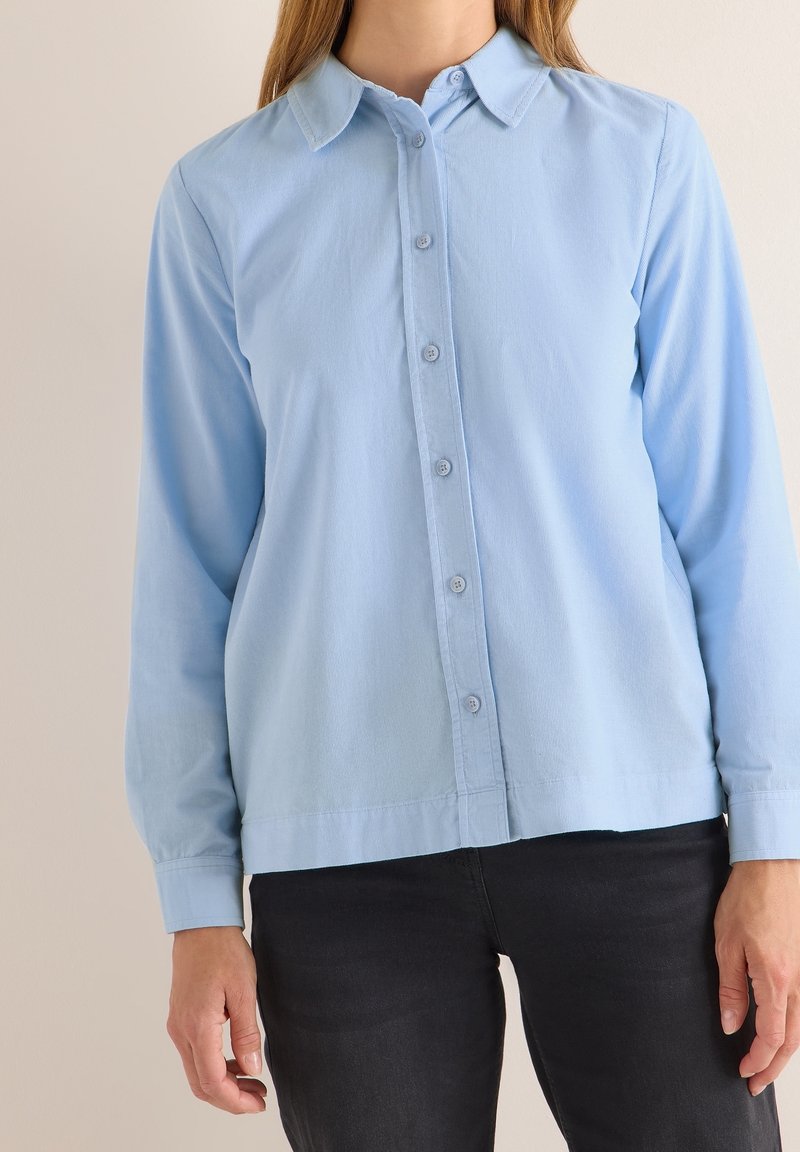 Femme portant une chemise bleu clair à manches longues boutonnée et un pantalon noir, debout devant un fond clair uni.