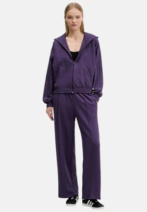 Giovane donna in piedi che indossa una felpa viola con cerniera e pantaloni a gamba larga abbinati, con scarpe da ginnastica nere e bianche.