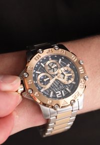 Montre chronographe à quartz avec un boîtier en acier inoxydable noir et doré, un cadran noir, des aiguilles luminescentes et un bracelet en maillons métalliques.