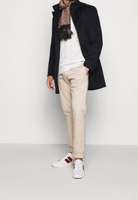 Cappotto nero, maglione bianco, pantaloni beige e sneakers bianche con strisce rosse. Sciarpa marrone a motivo. Texture morbide, vestibilità su misura.