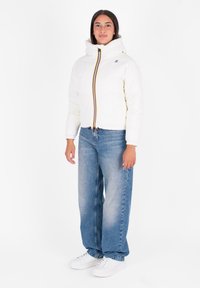 Witte pufferjas met een hoge kraag, voorzien van een kleurrijke rits en een trekkoord onderkant. Gecombineerd met lichtblauwe baggy jeans en witte sneakers.