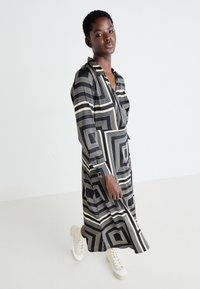 Abito geometrico bianconero con maniche lunghe, scollatura a V e una cintura in vita, abbinato a sneaker chiare.