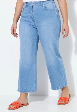 Person iført lyseblå vidde jeans og orange åbne flade sandaler, stående mod en hvid baggrund.