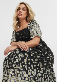 Svart klänning med vit och gul prästkragetryck. Har smockad byst och puffärmar. Mjuk tyg med en flödande kjol.