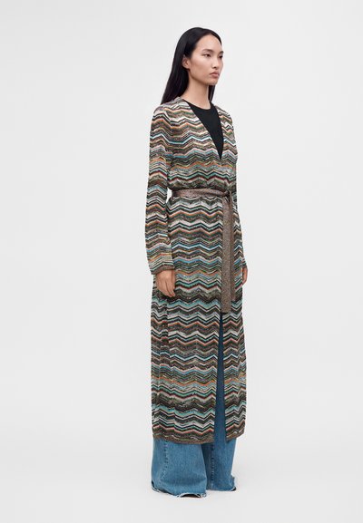 Femme debout portant un long cardigan multicolore à motif zigzag avec une ceinture sur un haut noir et un jean bleu à jambes larges.