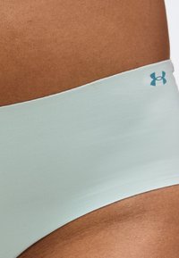 Κοντινή λήψη της ζώνης μέσης Under Armour σε ανοιχτό μπλε χρώμα πάνω σε σκουρόχρωμη μέση, που δείχνει το απαλό ύφασμα και το λογότυπο στη δεξιά πλευρά.
