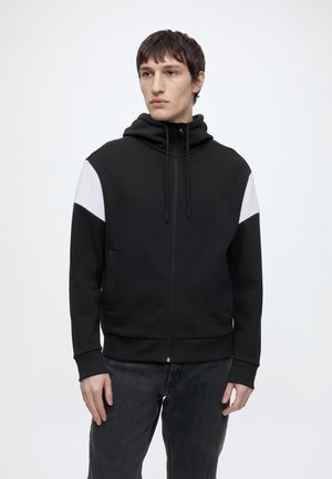 Jonge man met een zwarte hoodie met rits, witte schouderpanelen en donkere spijkerbroek, staand tegen een effen witte achtergrond.