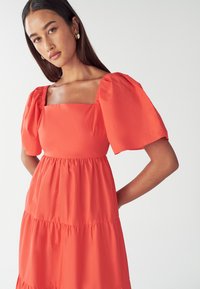 Sarkana kleita ar puff piedurknēm, kvadrātveida izgriezumu, daudzslāņu dizainu un gludu tekstūru. Iezīmē savāktus akcentus plecos un jostasvietā.