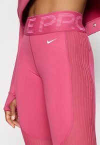 Różowe legginsy sportowe z szerokim pasem w talii, na którym widnieje tekst "PRO" w jaśniejszym odcieniu, małe białe logo Nike i teksturowane panele boczne.