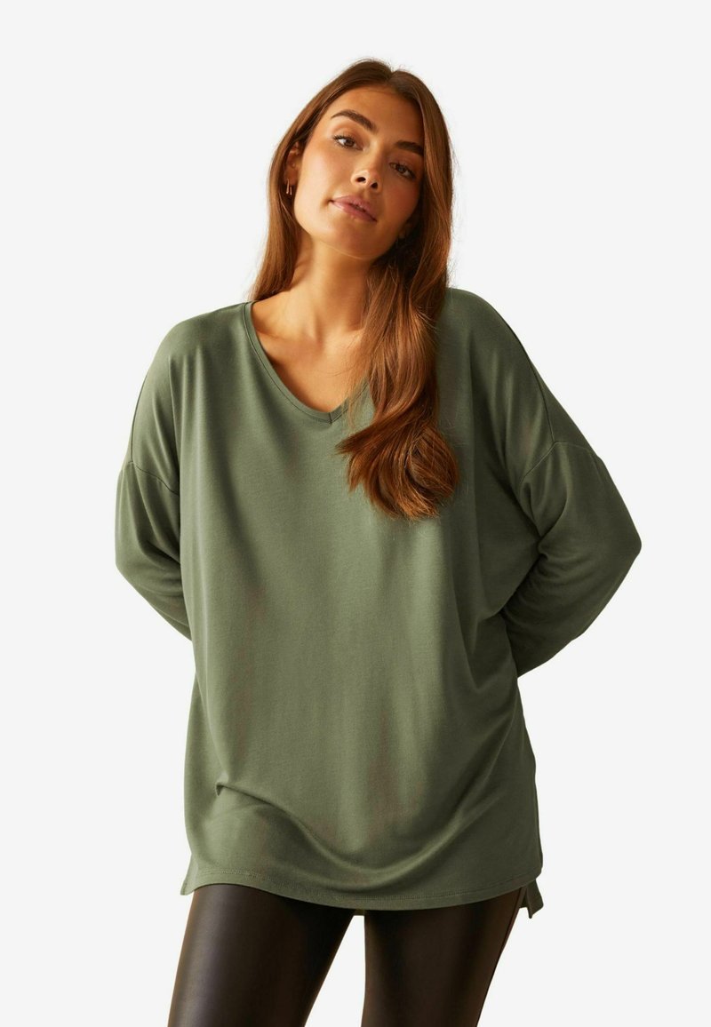 Grünes, übergroßes V-Ausschnitt-Shirt aus weichem Material, mit lockeren Ärmeln und abgerundetem Saum, kombiniert mit schwarzen Leggings.