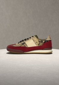 Αθλητικό παπούτσι με κόκκινο σουέντ, πάνελ από δέρμα σε μπεζ και σχέδιο φιδιού, μαύρα κορδόνια και μπεζ σόλα σε ανοιχτό γκρι επιφάνεια με γκρι φόντο.