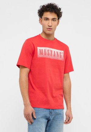 Young man with curly hair wearing a red Mustang logo t-shirt and blue jeans standing against a plain light background.