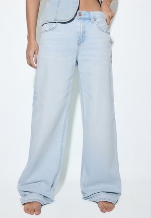Jean large bleu clair porté pieds nus, associé à un gilet en denim clair court, montré de la taille aux pieds.