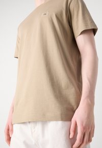 Beige bomullströja med rund halsringning, korta ärmar och en liten logotagg på framsidan. Slät textur, avslappnad passform.