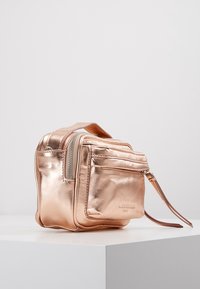 Roséguld metallic handväska, kompakt fyrkantig form, slät textur, med en sidficka med dragkedja och ett handledsband för enkel bärning.