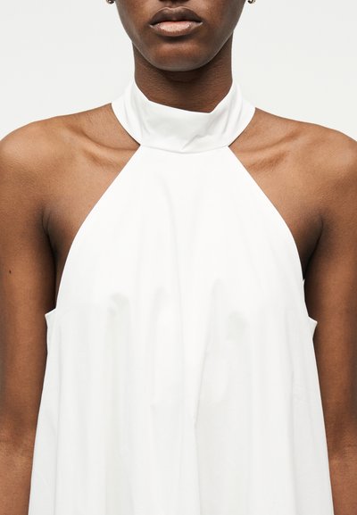 Gros plan d'une personne portant un haut blanc sans manches à col montant, montrant les épaules et le haut de la poitrine.