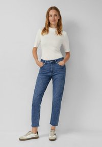 Witte korte mouwen top, blauwe hooggeripte jeans en beige sneakers met zwarte accenten. Model staat met de handen in de zakken op een grijze achtergrond.