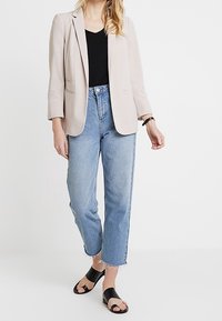 Blazer beige clair, haut noir, jean droit bleu clair et sandales noires. Le blazer présente des revers crantés et des manches trois-quarts.