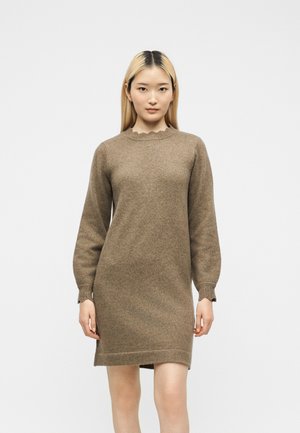 Femme aux cheveux blonds et raides, portant une robe tricotée marron à manches longues et longueur genou, debout devant un fond blanc uni.