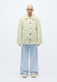 Faux fur krämjacka med krage och fem svarta knappar, i kombination med ljusblå vida jeans och beige skor.