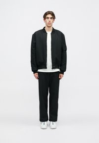 Μαύρο μπουφάν bomber με φερμουάρ μπροστά και πλευρική τσέπη, φορεμένο πάνω από ένα λευκό πουκάμισο και συνδυασμένο με μαύρο παντελόνι και λευκά αθλητικά παπούτσια.