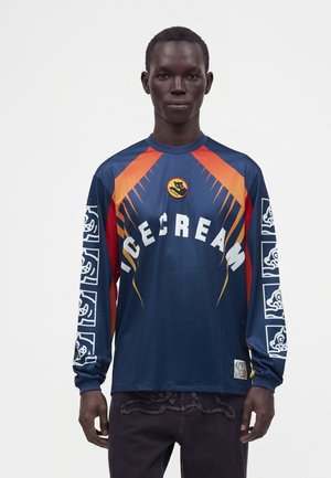 Mand iført en blå langærmet trøje med orange accenter, hvid tekst "ICECREAM," og grafiske designs på ærmerne mod en ensfarvet baggrund.