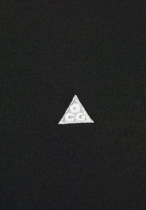 Svart tyg med en liten, broderad vit triangel-logotyp med bokstäverna "G" och "C" på. Texturen är slät och något elastisk.