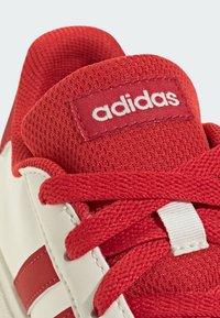 Crvena i bijela atletska cipela s mrežastim gornjim dijelom, teksturiranim crvenim vezicama i crvenom adidas oznakom. Ima bijele kožne detalje i crvene pruge.
