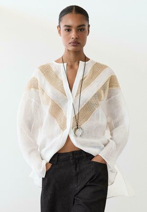 Frau trägt eine transparente weiße Bluse mit beigen gewebten Chevron-Details, schwarze Hose und eine lange schwarze Halskette mit einem silbernen Hufeisen-Anhänger.