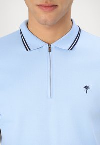 Světle modré polo tričko s límcem na zip, se středně modrými pruhy a malým vyšitým logem na levé straně. Vyrobeno z texturované látky.