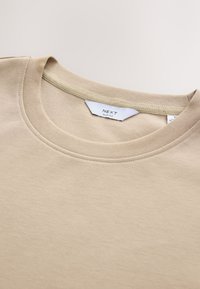 T-shirt beige coupe slim en coton doux avec une texture lisse. Encolure ronde avec un détail de couture subtil et une étiquette affichant les informations de la marque.