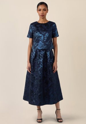Frau trägt ein marineblaues, florales Jacquard-Kurzarmshirt mit passendem, hoch tailliertem, plissiertem Midirock und schwarzen, knöchelhohen Riemchenabsätzen.