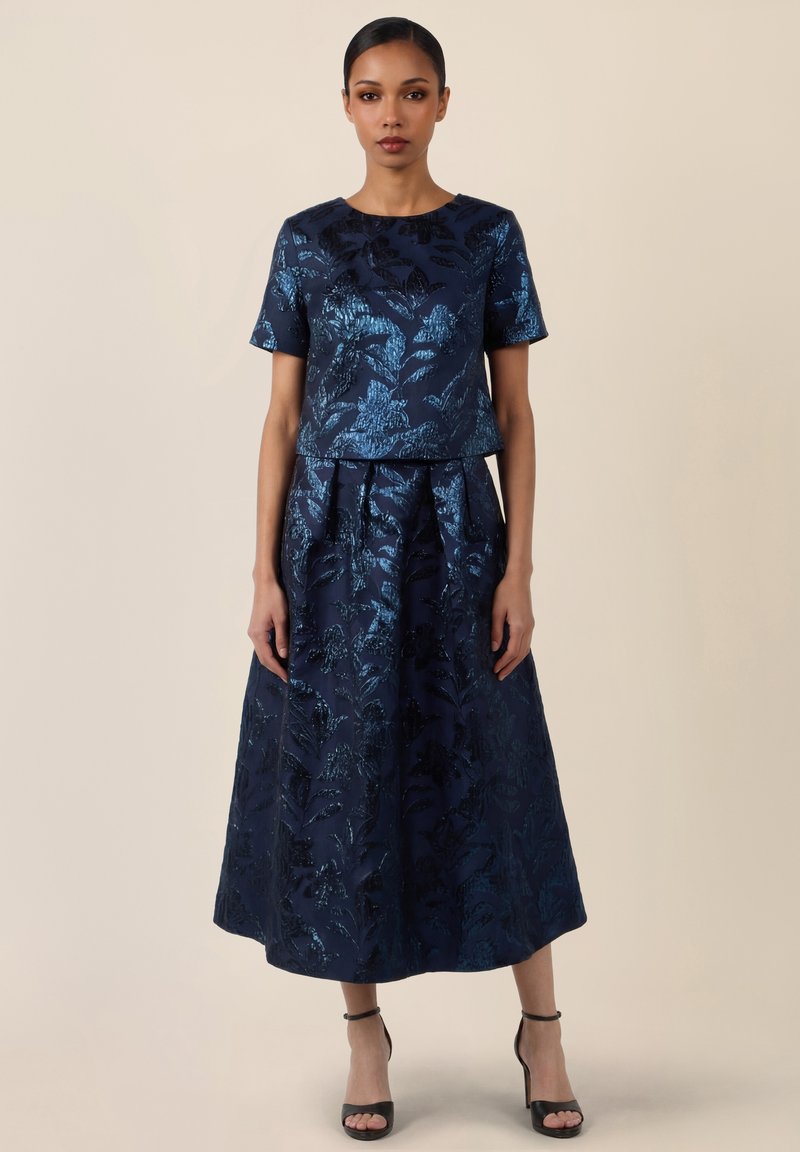 Femme portant un haut jacquard à manches courtes bleu marine à motif floral, assorti à une jupe midi plissée taille haute, et des escarpins noirs à bride à la cheville.