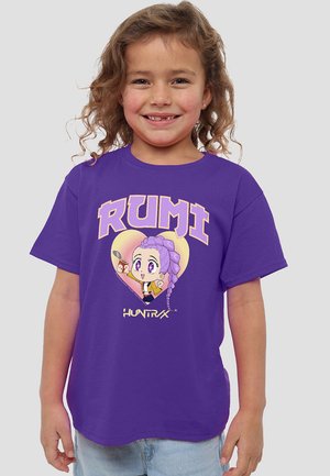 Smiling child with curly hair wearing a purple T-shirt featuring a cartoon character with purple hair inside a yellow heart and text "RUMI" and "HUNTRX".