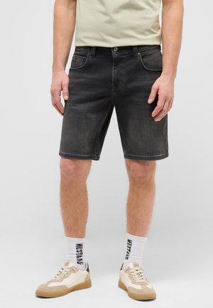 Mann trägt schwarze Denim-Shorts, weiße Sportsocken mit "MUSTANG"-Schriftzug und beige-weiße Sneaker, steht mit entspannt an den Seiten hängenden Armen.