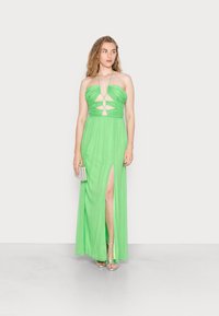 Vestido maxi verde sem alças com detalhes recortados, tecido fluido e uma racha até a coxa; combinado com sandálias prateadas e uma pequena clutch.