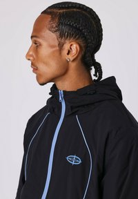 Chaqueta negra con capucha y acentos en azul claro, con cremallera, que presenta un logo en el pecho y un tejido ligero y texturizado.