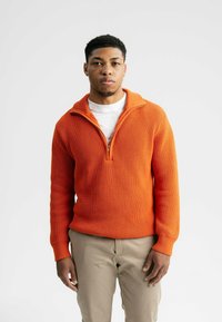 Oranger rippenstrukturierter Pullover mit einem halben Reißverschlusskragen, getragen über einem weißen T-Shirt, kombiniert mit beigen Chinos.