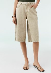 Beige denimshorts med løs pasform, midt på læggen, og store lommer. Sammen med sorte og beige sandaler med remme.