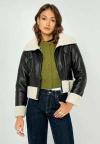 Giacca cropped nera in pelle con colletto e polsini in finta shearling bianca, tasche con zip, indossata su un maglione verde a coste e jeans blu.