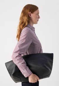 Borsa tote oversize in pelle nera tenuta nella mano destra, caratterizzata da una texture liscia e una forma geometrica. Modella con indosso una camicia a righe.