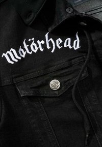 Svart denimjacka med vit broderad text "Motörhead" och en silverfärgad dödskalleknapp på bröstfickan. Texturerad tyg med en avslappnad design.