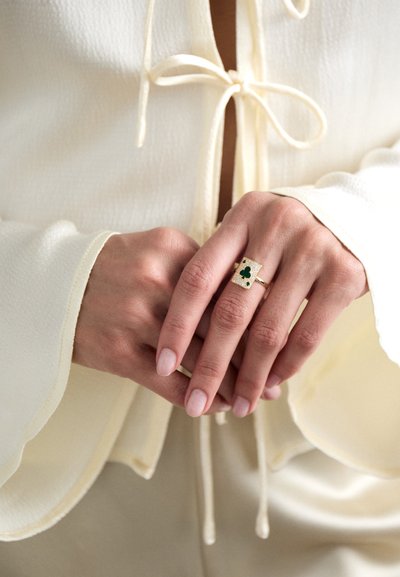 Anillo de oro con un diseño de trébol de esmalte verde en un ajuste rectangular texturizado, llevado en una mano con esmalte de uñas en tonos neutros.