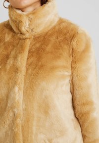 Fuskpälsjacka i ljusbeige med hög krage. Mjuk textur, slät yta och sömlös design med ett varmt, plyschigt utseende.