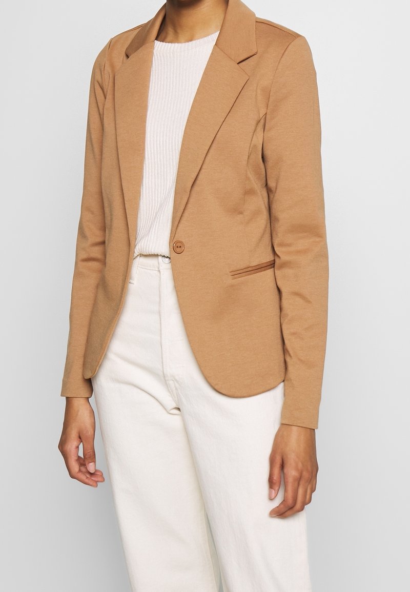 Světle hnědý blazer vyrobený z měkké látky, s jedním knoflíkem, zářezovými klopami a slim fit střihem. Nositý přes proužkovaný krémový top a bílé kalhoty.